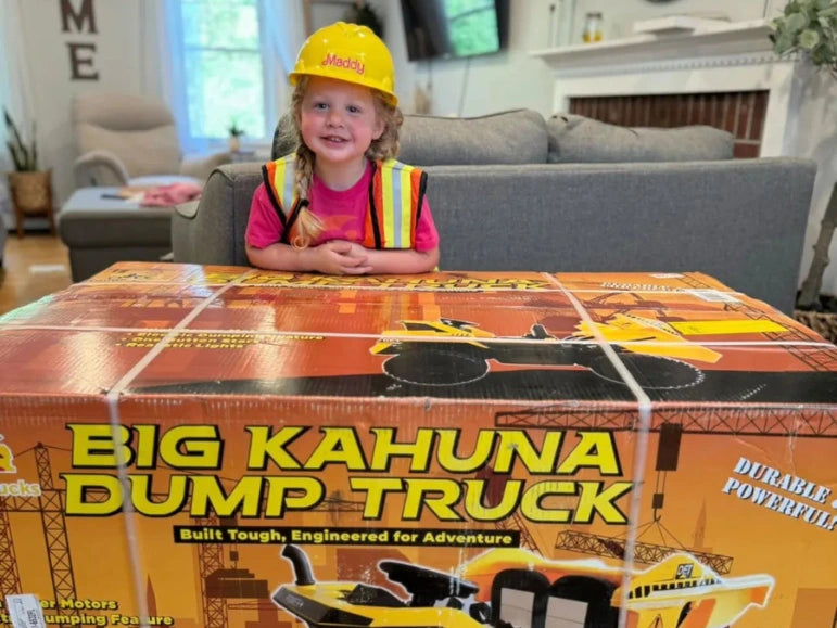 Girl wearing hard hat with Maddy written on it is looking at camera and smiling while standing behind the box of Big Kahuna Dump Truck by Tough Trucks For Kids